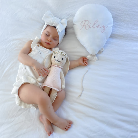 Newborn baby in a white outfit holding a plush balloon, lying on a textured white surface with a heart-shaped pillow.