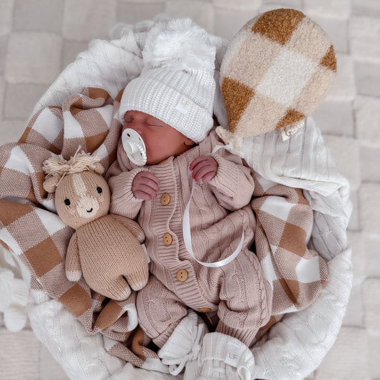 Newborn baby in a basket with checkered blanket, knitted toy, and knitted hat on a textured surface.